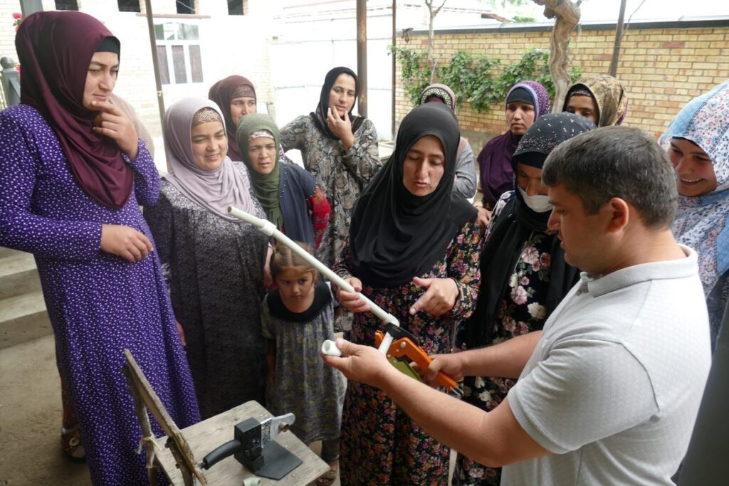 A group of women in headscarves are gathered around a man holding a water pipe that is training them on how to use community plumbing systems.