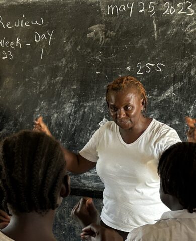 A woman speaks to children in front of a blacboard.