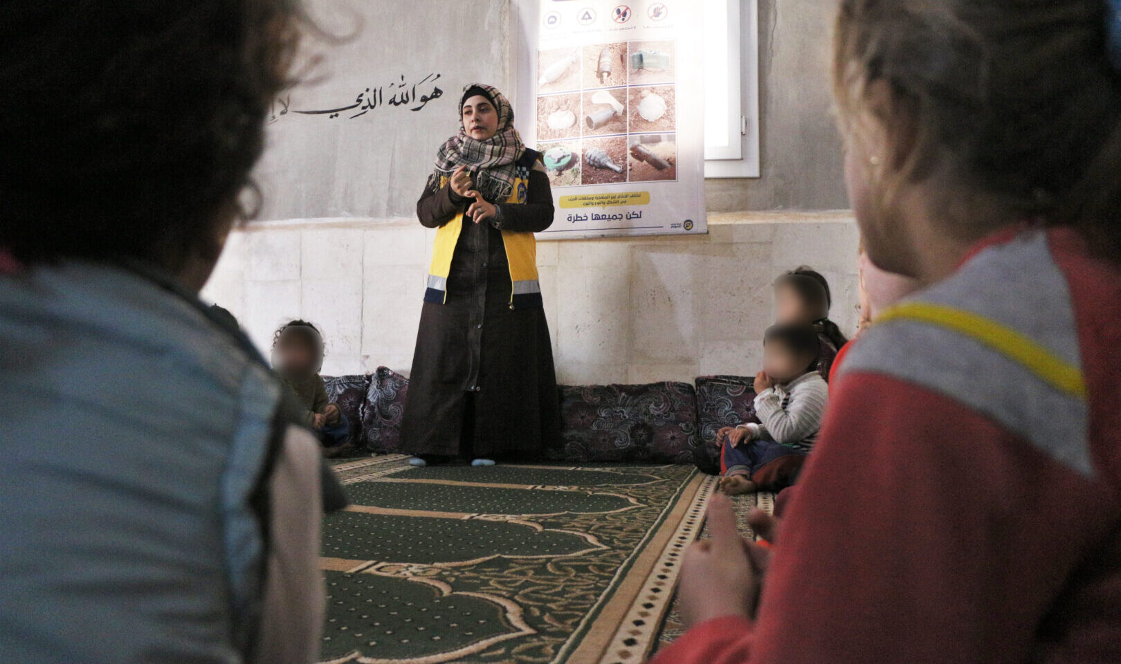 A woman in a headscarf speaks to children sitting on a floor.