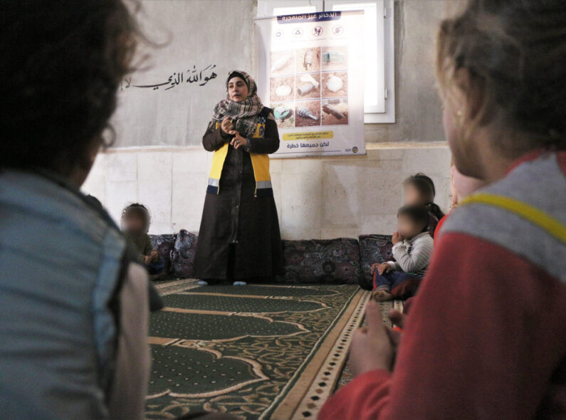A woman in a headscarf speaks to children sitting on a floor.