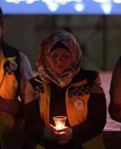 Three White Helmets volunteers stand holding lit candles in their hands, with heads bowed