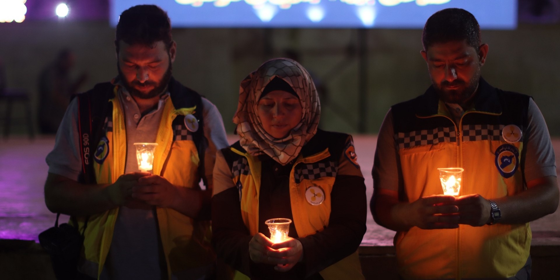 Three White Helmets volunteers stand holding lit candles in their hands, with heads bowed