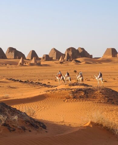 A view across the desert in Sudan, with four people riding camels in the foreground and the Pyramids of Meroë in the background
