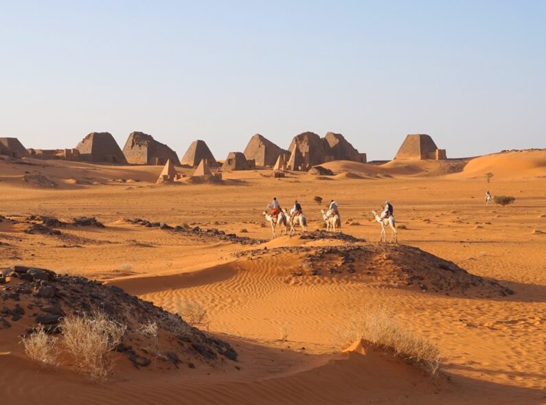 A view across the desert in Sudan, with four people riding camels in the foreground and the Pyramids of Meroë in the background