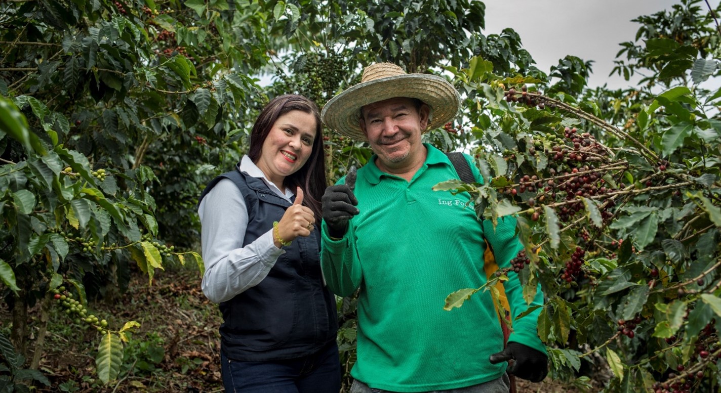 A woman in a black vest and a man in a green shirt pose with their thumbs up in front of a field, surrounded by coffee plants.