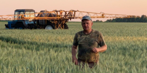 A man stands in a field of green crops, with a large piece of machinery behind him