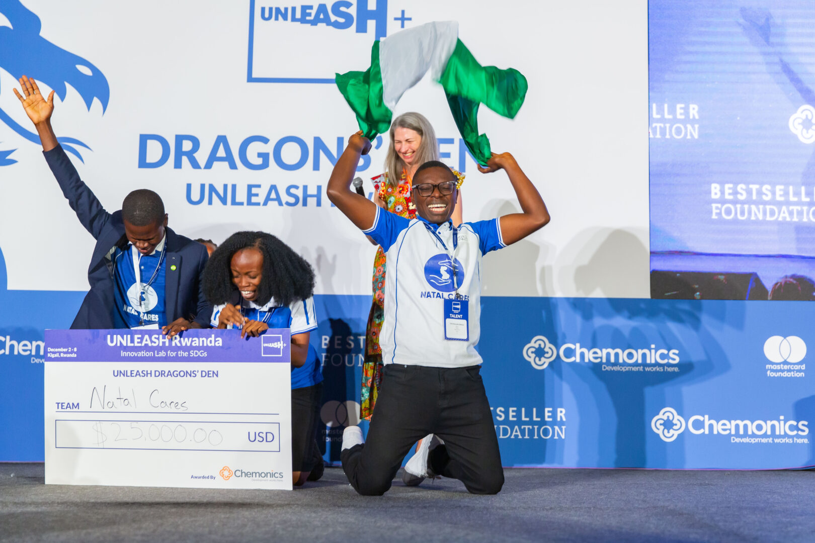 A man excitedly jumps with the Nigerian flag. Two people kneel next to him with a giant check. A man puts his hand in the air with excitement. 