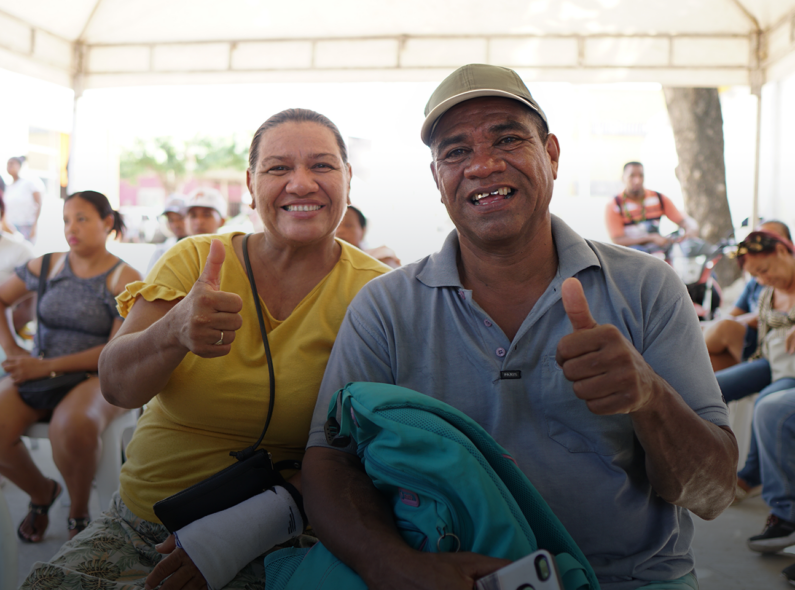 A man in a blue shirt and khaki baseball cap and a woman in a yellow shirt smiling and giving a thumbs up to the camera.