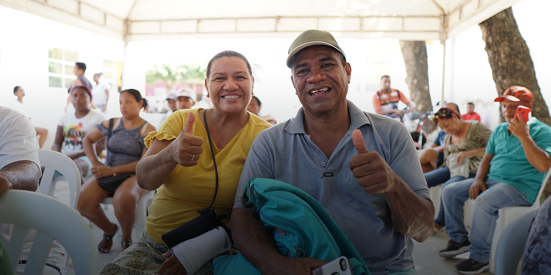 A man in a blue shirt and khaki baseball cap and a woman in a yellow shirt smiling and giving a thumbs up to the camera.