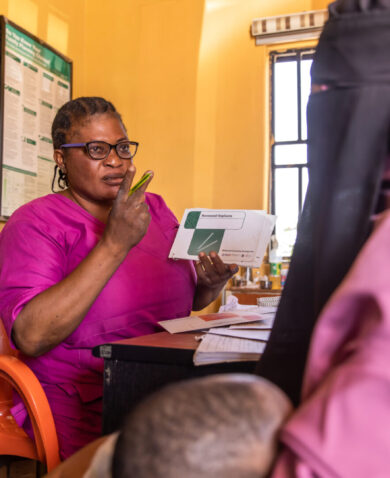 A Nigerian nurse wearing bright pink and glasses speaks to another woman holding a child behind a desk.