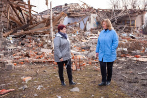 Two women, Halyna Minayeva and a colleague, in front of a building's destruction.