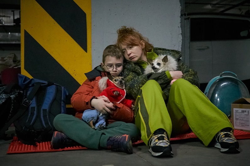A young boy and a woman sit on the floor of a parking garage, leaning against a pillar. Both are holding small dogs, and there are bags on the floor beside them.