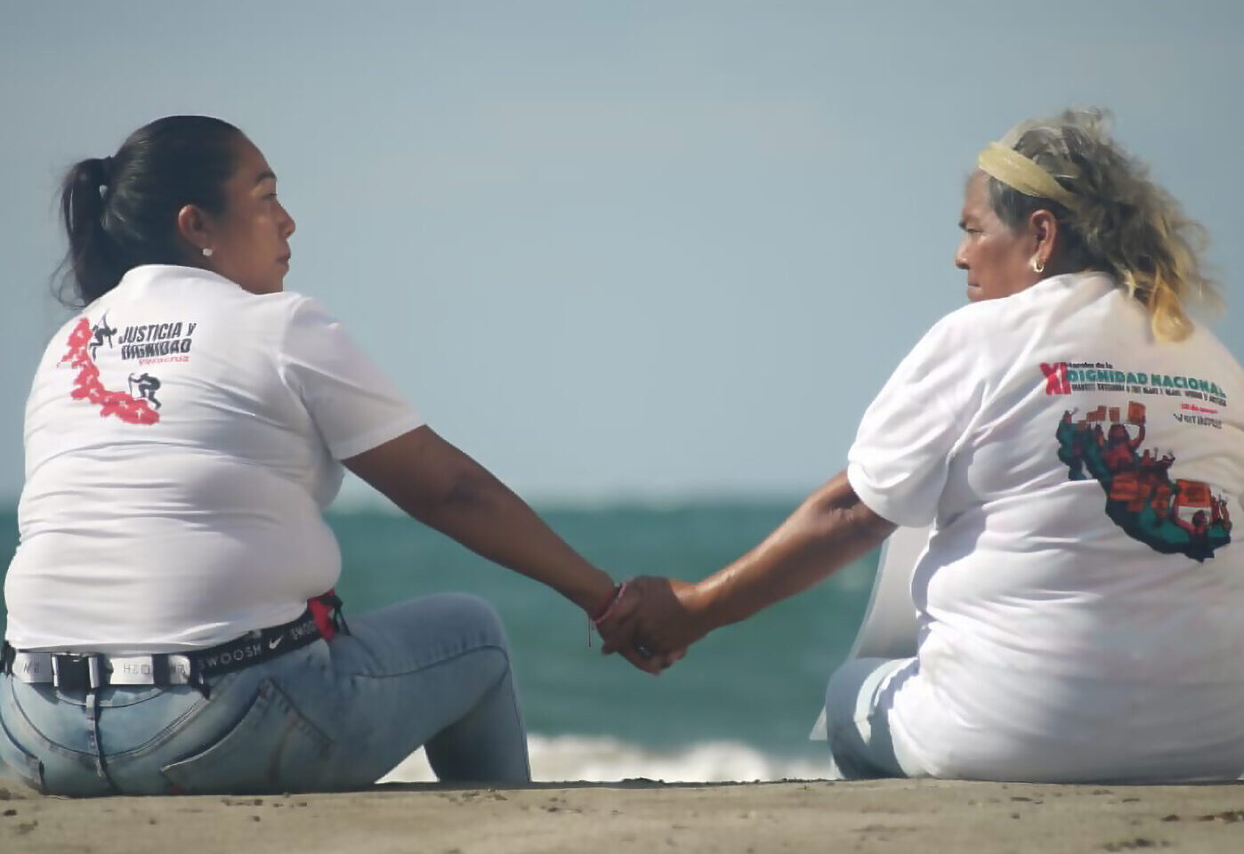 Two women in white tshirts holding hands in front of the ocean.
