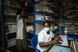 A health worker at a facility in DRC uses InfoMED to track stock level data of health commodities while another places boxes of commodities in a shelf in the back. Credit: Arlette Bashizi.
