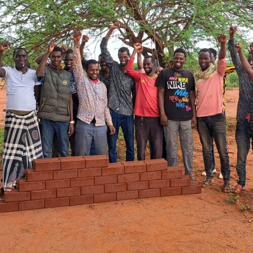 A group of young men stand behind the start of a wall they are building using the bricks they have made