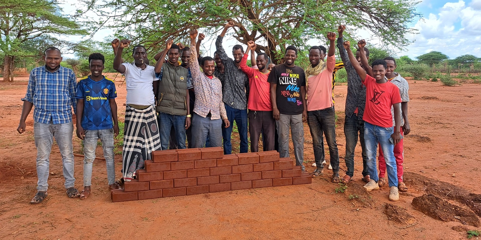 A group of young men stand behind the start of a wall they are building using the bricks they have made