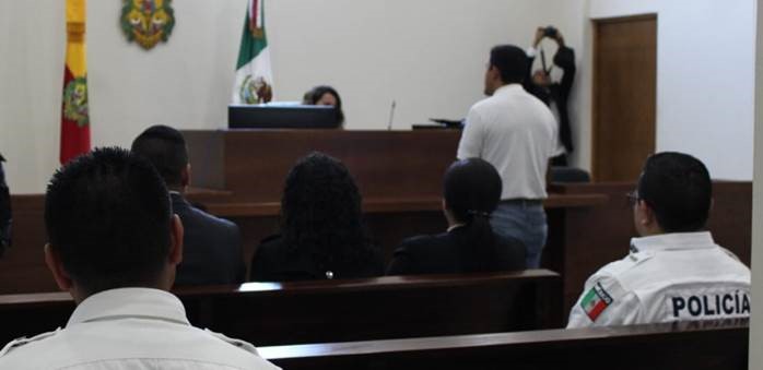 A court room with a man standing and speaking to the judge. A police officer's back is to the camera in the foreground