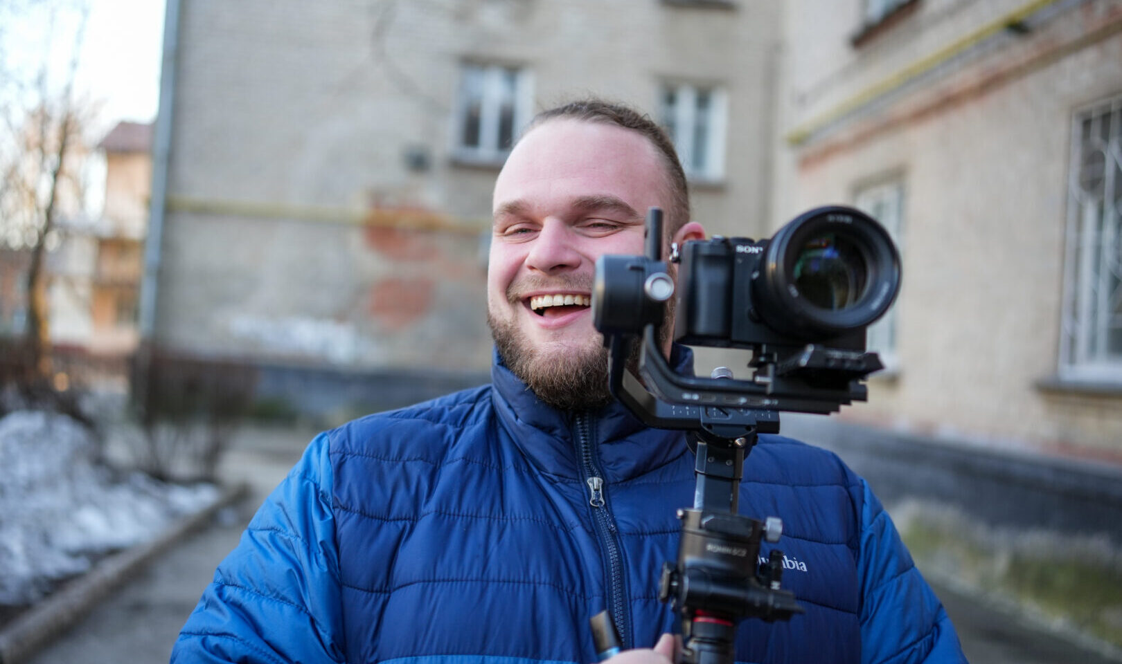 A man in a blue down jacket holds gleefully holds a camera on a stabilizer.