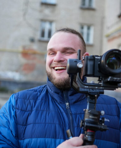 A man in a blue down jacket holds gleefully holds a camera on a stabilizer.