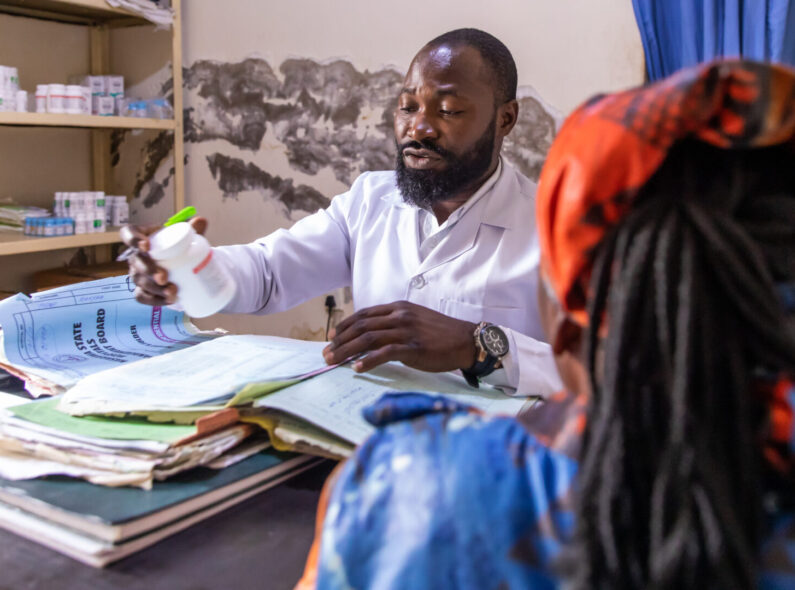 A health worker holds a bottle of HIV medicine while at his desk speaking to a woman patient