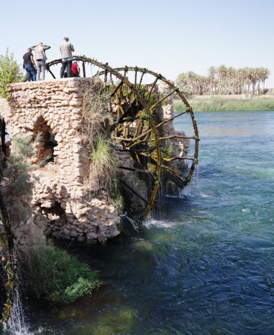 People stand alongside 2 water wheels in a river. A boat floats below.