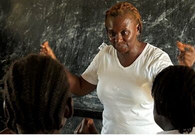 Two adults stand in front of a chalkboard dusted with chalk in front of a group of children. One adult, a woman with ginger braids in a white shirt, holds her hands longways to teach the children.