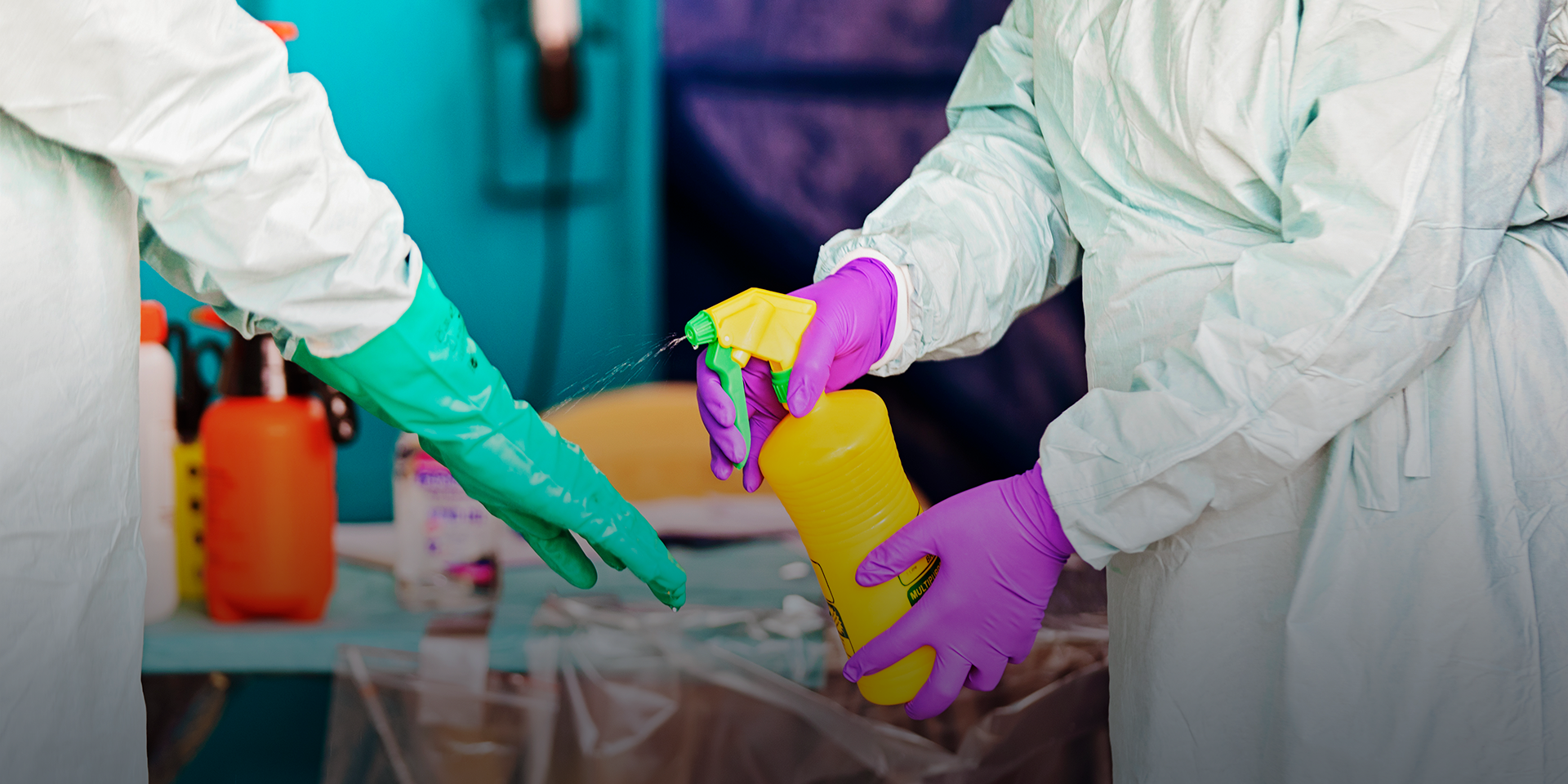 Purple gloved hands are spraying a liquid on a person's green-gloved hands