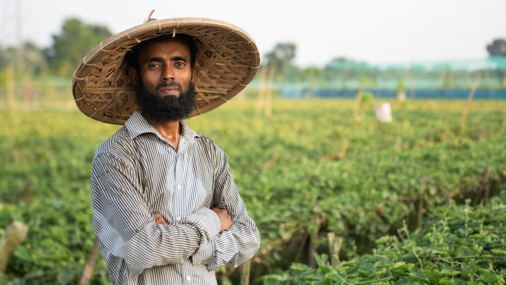 A man stands and looks at the camera, standing in front of a field of green crops.