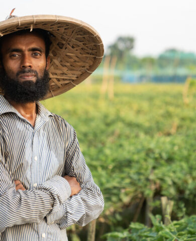 A man stands and looks at the camera, standing in front of a field of green crops.