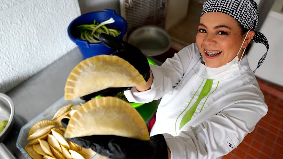 A woman in her chef's outfit holds up two empanadas to the camera