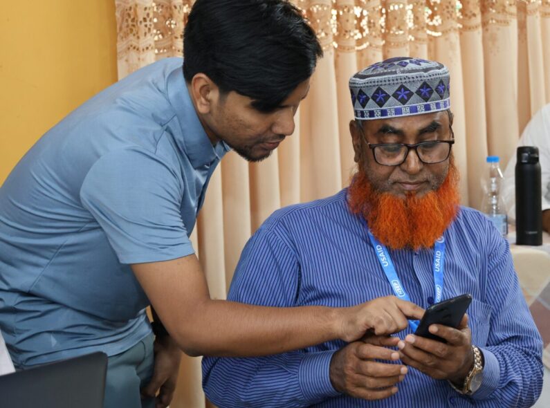 A young man leans over to show an older man how to navigate an online system on his phone