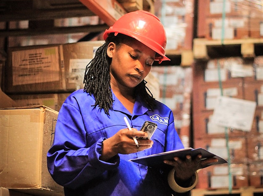 A woman wearing a hard hat uses her phone to scan a piece of paper inside a warehouse.